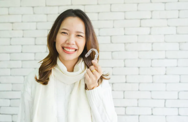 Smiling woman holding Invisalign clear aligner in front of a white brick wall, illustrating orthodontic treatment options.