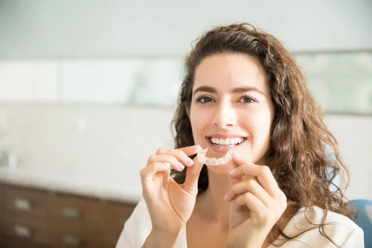 Woman smiling while holding Invisalign aligner in orthodontic office, representing effective orthodontic treatment options.