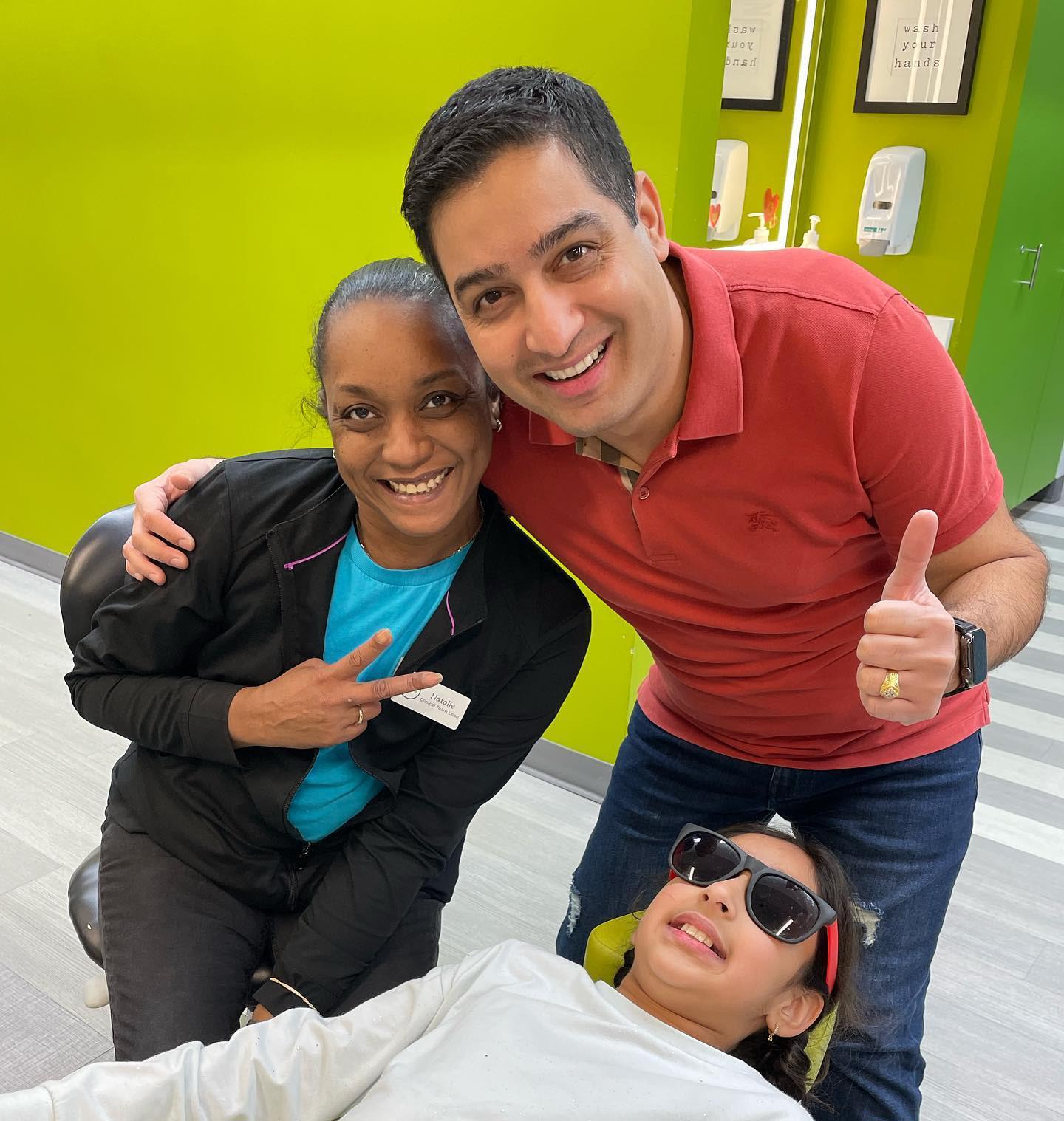 Pediatric dentist and staff posing with a young patient wearing sunglasses in a colorful dental office, promoting a positive dental experience for children.